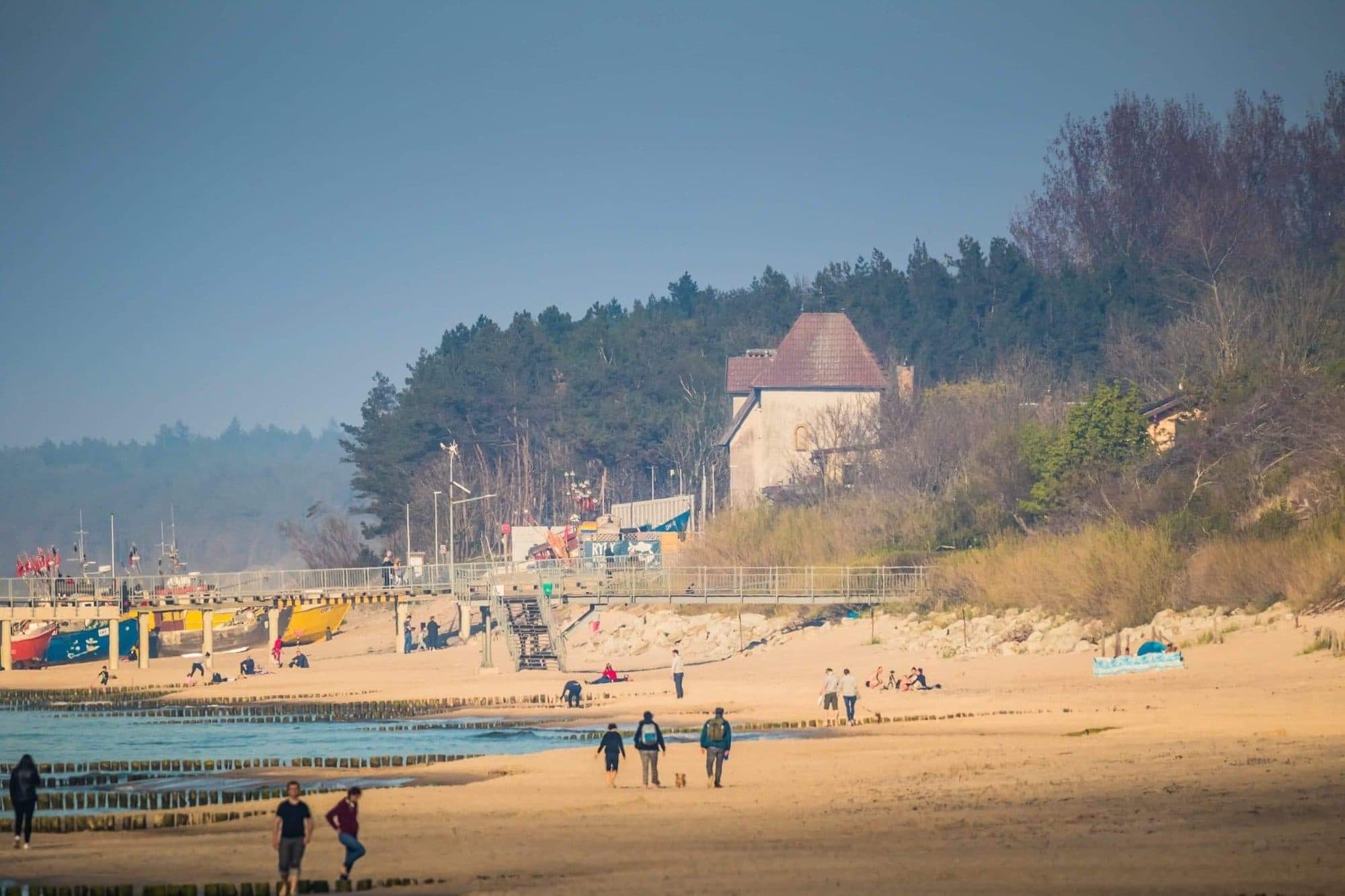Plaża w Sarbinowie ze spacerowiczami, falochronami i kutrami rybackimi przy pomoście